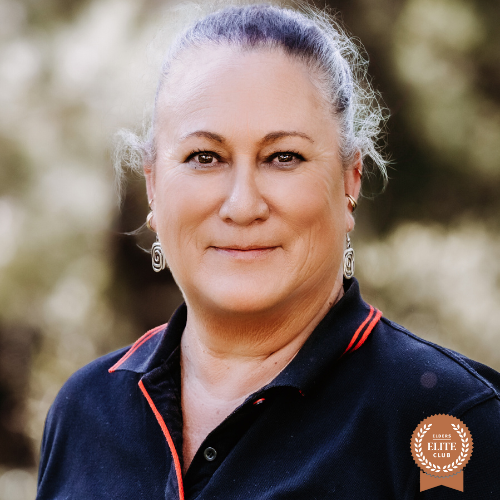 A woman with long gray hair styled in a ponytail, smiling warmly at the camera while wearing a black polo shirt with red accents.